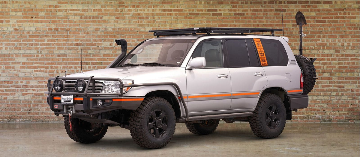 A silver Toyota 100 Series Land Cruiser SUV with custom off-road modifications, including a black roof rack, front bumper guard, snorkel, and spare tire on the rear, parked against a brick wall.