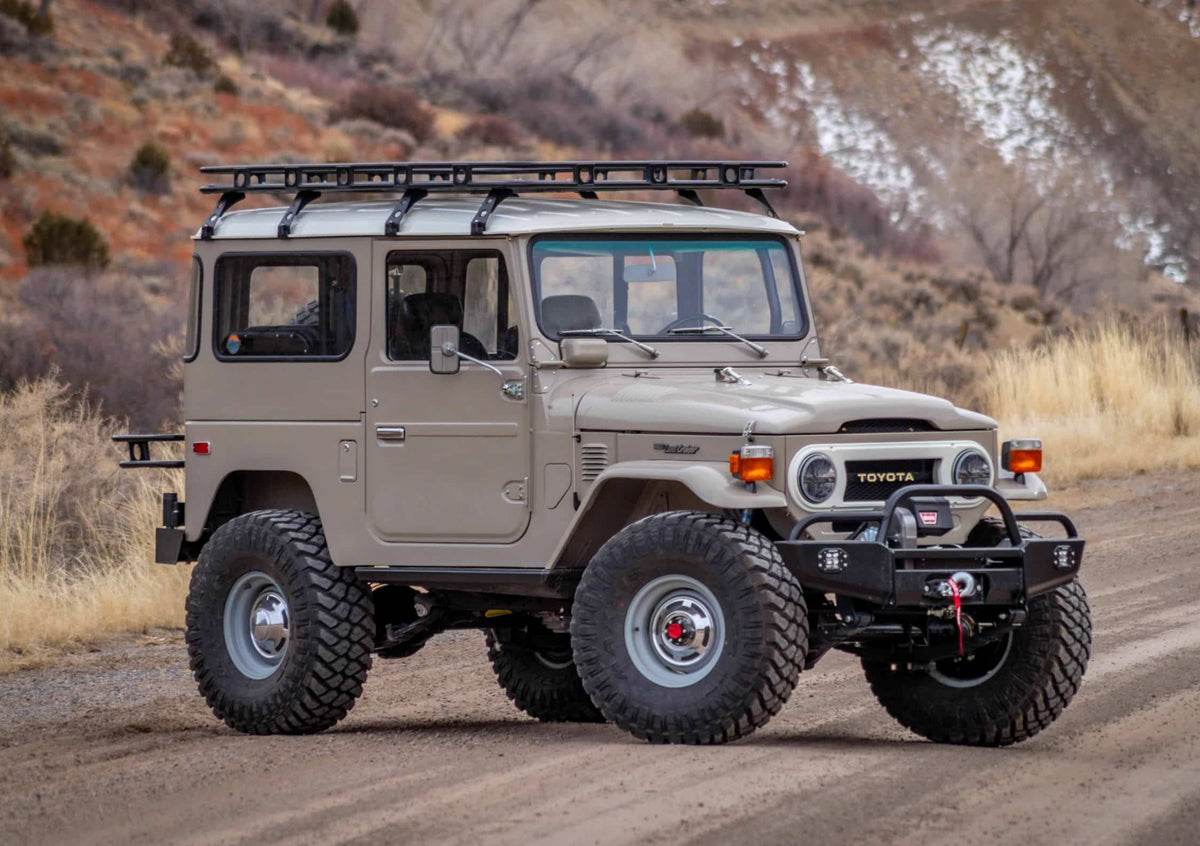 A beige Toyota FJ40 Land Cruiser off-road vehicle with a roof rack and heavy-duty front bumper, parked on a dirt road with dry grass and hills in the background.
