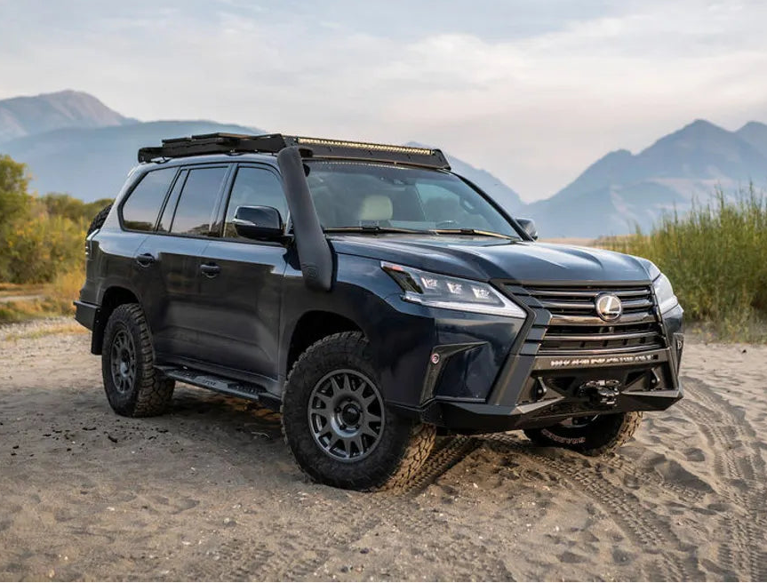 A dark gray Lexus LX 570 SUV with off-road modifications, including a roof rack, snorkel, and front bumper guard, parked on a sandy path with mountains in the background.