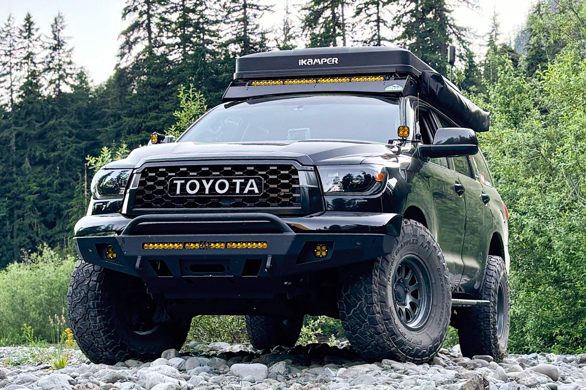 A black Toyota SEQUOIA 2nd gen with off-road modifications, including a roof-mounted and a front bumper light bar, parked on a rocky trail with a forest background.