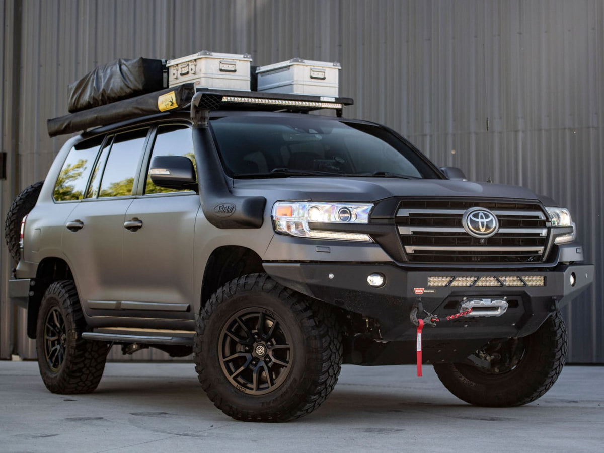 A customized dark gray Toyota 200 Series Land Cruiser SUV with off-road modifications, including a black front bumper with LED lights, a roof rack carrying two hard cases, and a spare tire on the rear, parked against a gray corrugated metal wall.