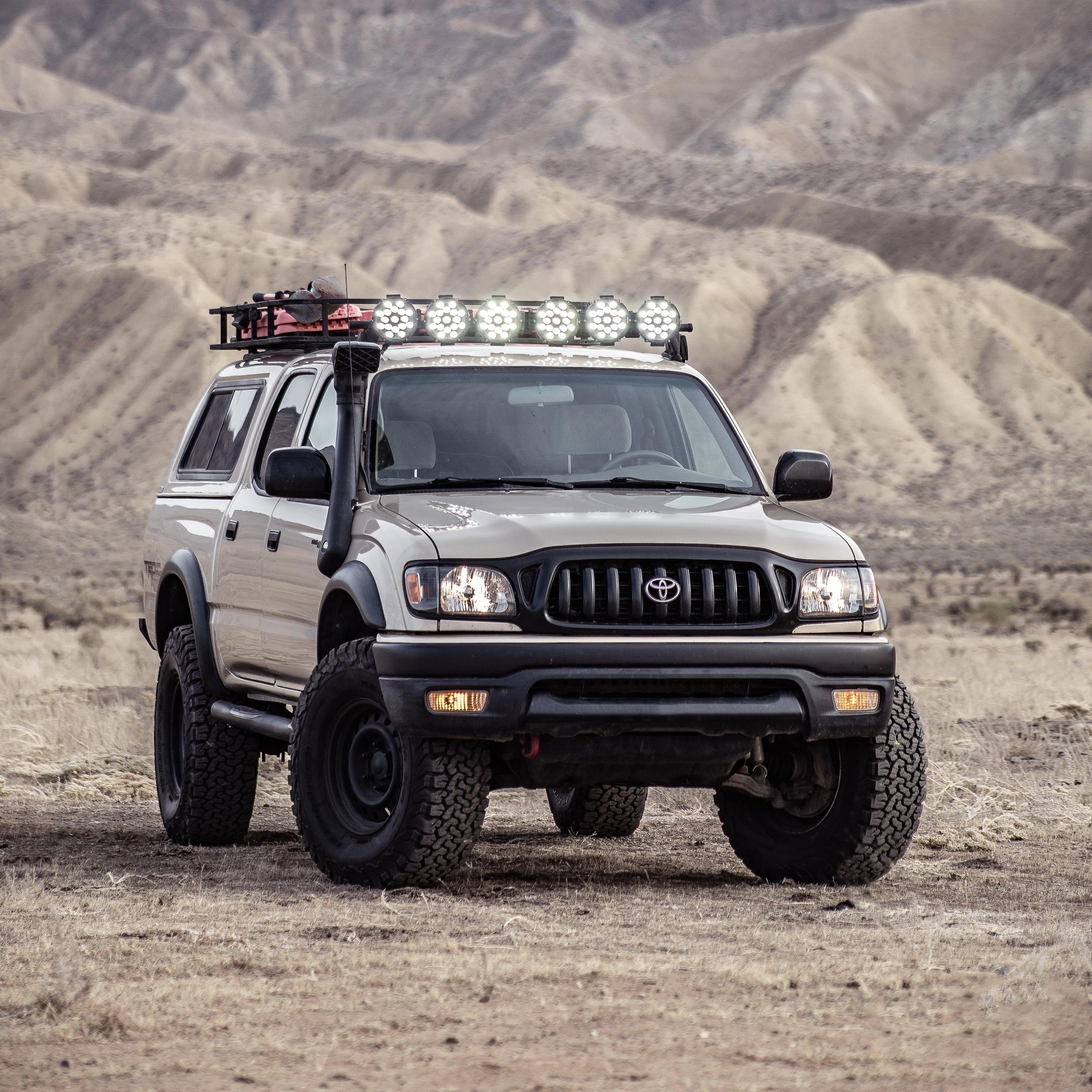 A lifted Toyota Tacoma pickup truck with off-road modifications, including a roof-mounted light bar, snorkel, and front bumper, parked in a desert landscape with rocky hills in the background.
