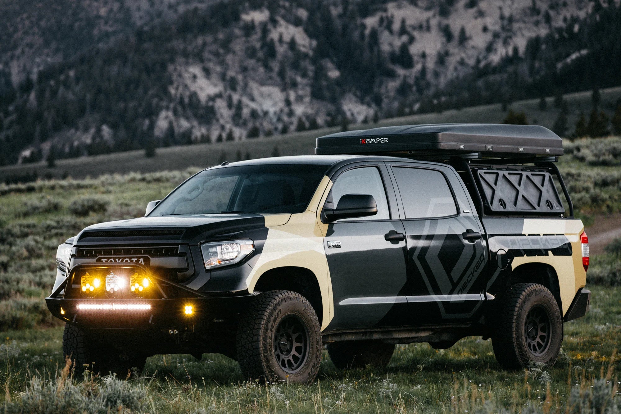 A customized black and beige Toyota Tundra pickup truck parked in a grassy mountain landscape. The truck features a black roof top tent, a front bull bar with bright amber lights, and geometric decals on the side.